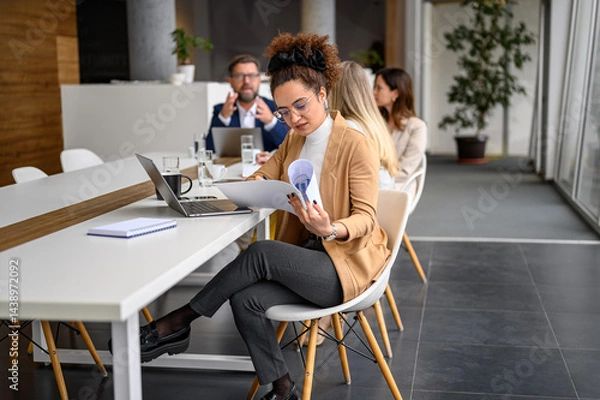 Fototapeta Businesswoman reviewing reports and working over laptop with colleagues in the background at modern office