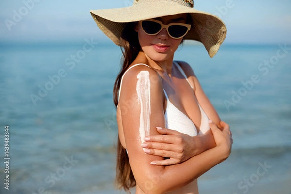 Fototapeta Woman applying sunscreen on her arm while enjoying a sunny beach day, wearing sunglasses and a wide-brimmed hat, embracing summer relaxation by the water.