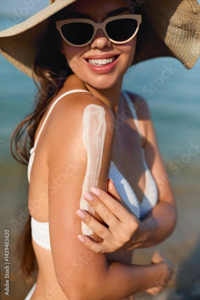 Fototapeta Woman applying sunscreen on her shoulder at the beach, enjoying the sun while wearing sunglasses and a wide-brimmed hat, promoting summer skincare and sun safety.