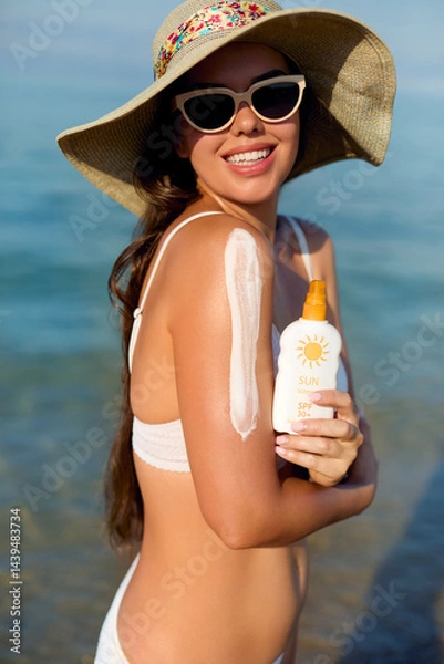 Fototapeta A beautiful young woman applying sun cream to her shoulder. Girl wears stylish sunglasses and enjoys the sun on a warm summer day