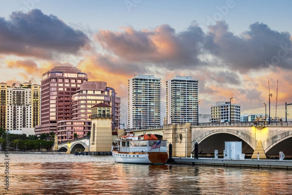Fototapeta Sunset view of West Palm Beach, Florida, showing a colorful sky, modern city skyline, yachts at the marina, historic bridge, and calm water reflections. Perfect for travel, cityscape, and architecture