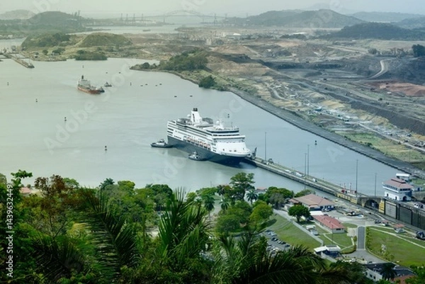 Fototapeta Aerial view of the Panama Canal with a cruise ship entering a lock chamber, surrounded by tropical vegetation, construction terrain, and distant infrastructure across the channel.