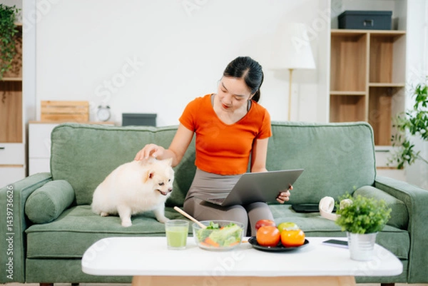 Fototapeta Woman relaxing on sofa at home with dog reading digital tablet in bright modern living room, joyful pet