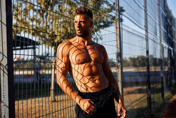 Obraz Muscular man with a defined six-pack leaning against a wire mesh fence, casting interesting shadows on his body