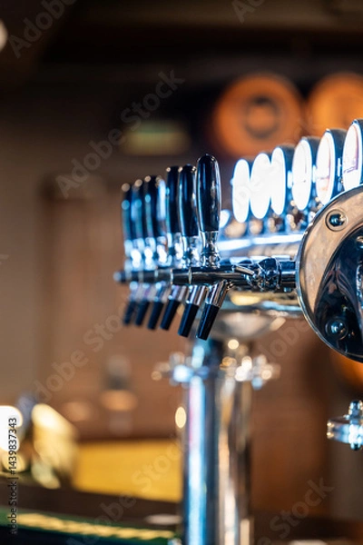 Fototapeta A close-up of a shiny beer tap system with black handles, gleaming under soft lighting in a bar setting. The blurred background suggests a cozy, inviting atmosphere for social gatherings.