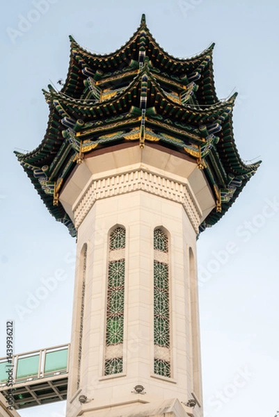 Fototapeta Minaret Tower of a Mosque in Sanya, Hainan, China with a green, blue, and gold roofline. The tower has two arched windows with intricate latticework and a bridge connecting to the tower.