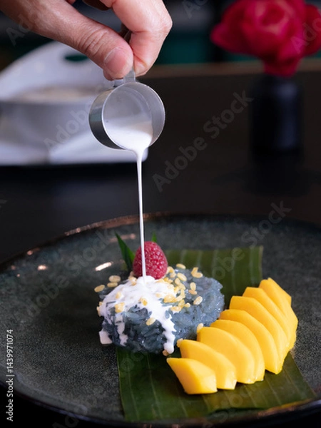 Fototapeta A plate of ripe mango with sticky rice and coconut milk sauce pouring on it, various sweet Thai desserts in background with Thai style plate decorations such as green banana leaves 
