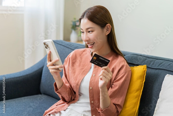Fototapeta Bill pay, shopping order online internet, cute asian young woman hand holding credit card and mobile smart phone on sofa at home, female using banking service to secure payment with purchase on store.