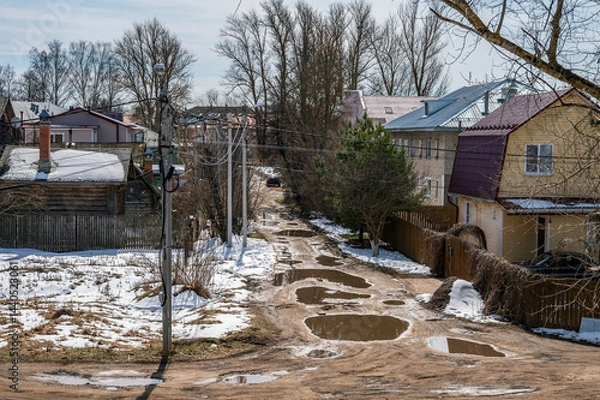 Fototapeta A dirt road on the street of a large village with large puddles in spring.