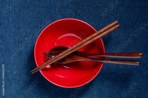 Fototapeta A pair of wooden chopsticks, fork and spoon lying across an empty red soup bowl on blue fabric background, close up, top view