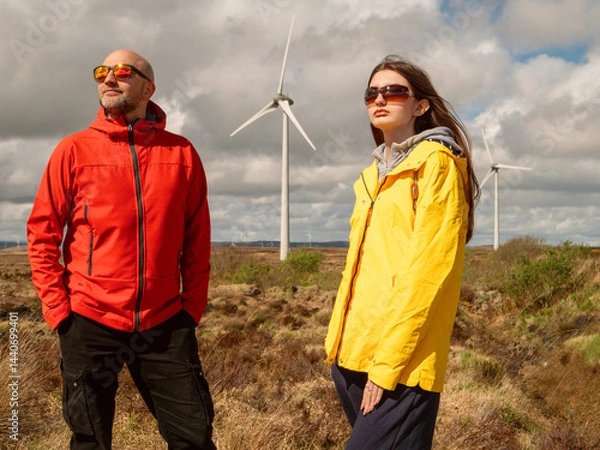 Fototapeta Father in red jacket and daughter in yellow enjoy warm sunny day. Wind turbine and huge wild field and cloudy sky in the background. Divorced man day with his daughter concept. Outdoor activity.