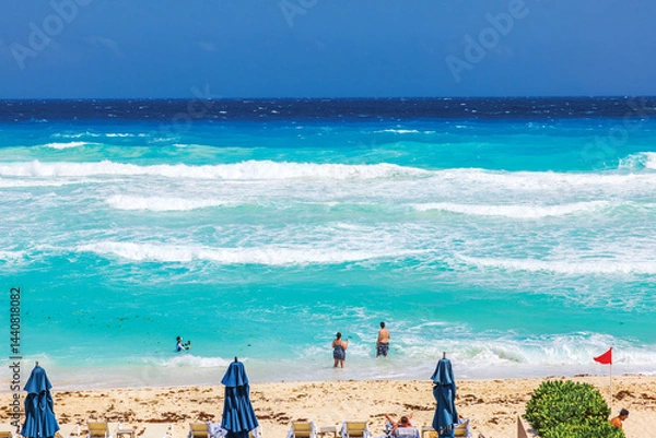 Fototapeta Tourists swimming in turquoise water of Caribbean Sea with waves and red flag warning on beach. Mexico. Cancun.