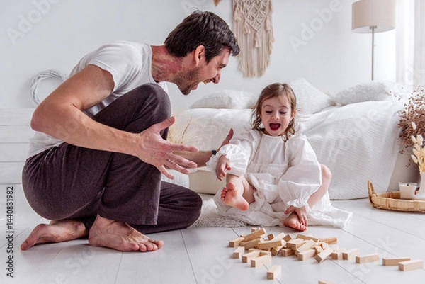 Fototapeta Dad and his little daughter sitting on the floor, playing joyfully with colorful wooden blocks, sharing laughter and creating cherished memories in their cozy bedroom