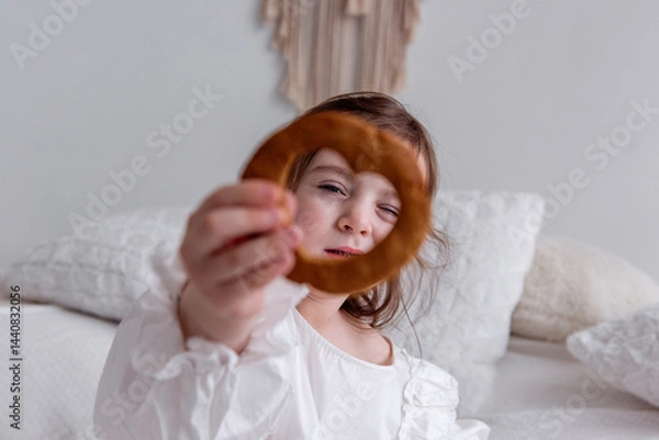 Fototapeta Little girl in a white dress sitting on a cozy bed, holding a heart-shaped cookie in front of her eye while smiling and winking playfully, radiating joy and innocence