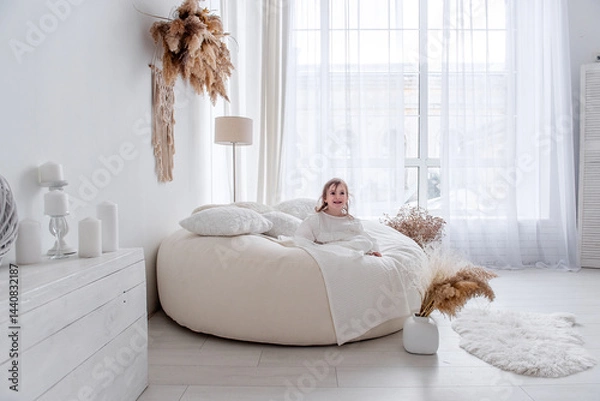Fototapeta Young girl enjoys a relaxing moment on a comfortable beanbag chair in a bright, bohemian-chic living room, surrounded by stylish decor and natural light streaming through a large window