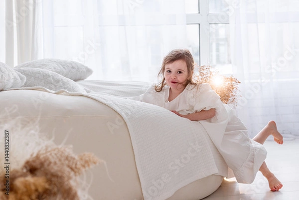 Fototapeta Cute little girl wearing a white dress joyfully playing on a bed in a bright white bedroom, embracing the cheerful atmosphere of a playful morning at home