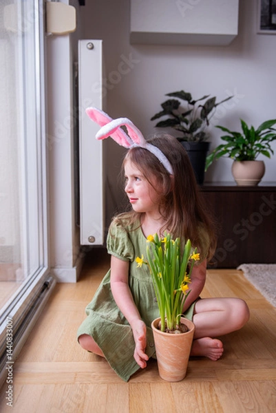 Fototapeta Cute child wearing Easter bunny ears headband sitting on the floor next to the window holding a pot of yellow daffodils waiting for Easter bunny
