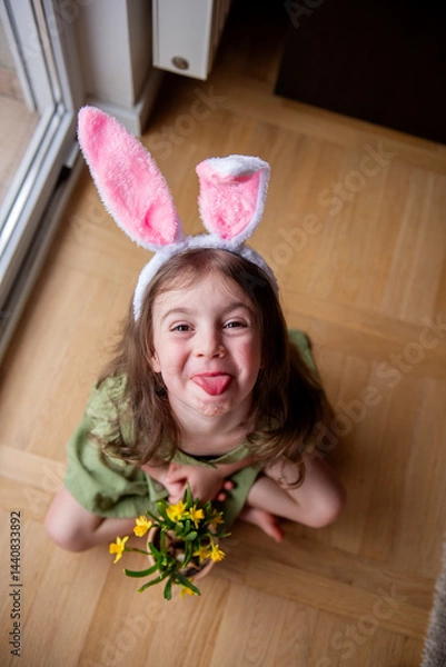 Fototapeta Little girl wearing bunny ears sits on the floor with her legs crossed, playfully sticking her tongue out while holding a pot filled with cheerful yellow flowers