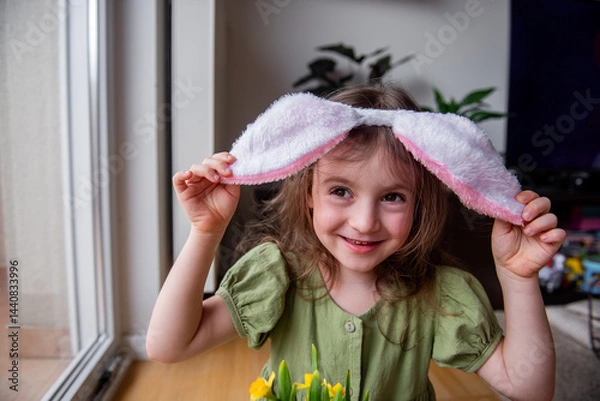 Fototapeta Little girl wearing festive Easter bunny ears, joyfully holding bright daffodils and smiling at home, radiating happiness and innocence in a playful indoor setting