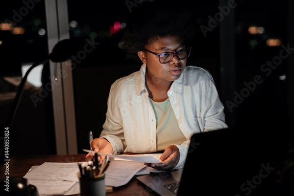 Fototapeta Young African businesswoman working on a laptop and paperwork in the evening