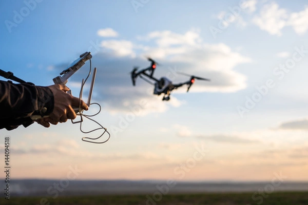 Fototapeta Demonstration of .copter Man controls quadrocopter flight. Flying the copter over a field over sunset sky. Remote control in a man's hands.