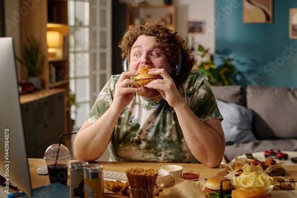 Fototapeta Person with curly hair enjoying a large sandwich in a cozy, cluttered room filled with various snacks and drinks, using device, and sitting at desk with computer screen visible