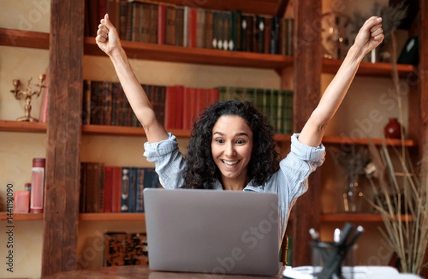 Fototapeta Yes, I'm A Winner. Excited emotional ecstatic lady celebrating success, victory or great news sitting at table, using personal computer, screaming yeah and raising hands up. Smiling happy woman in joy