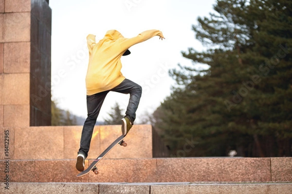 Fototapeta The teenager in a sweatshirt and a cap jumps with a board in the city against the backdrop of the urban sunset light. Photo of extreme