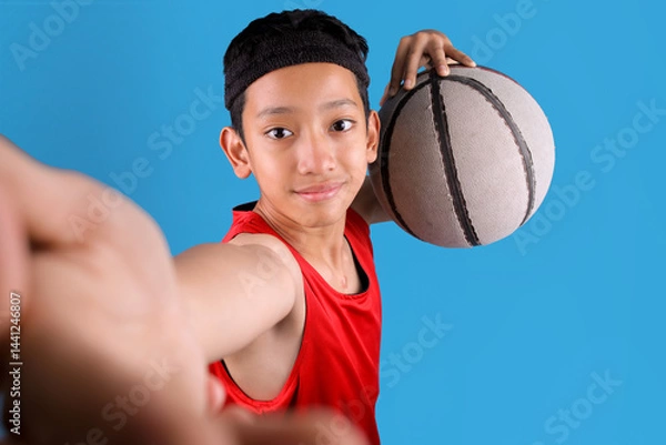 Obraz Portrait of smiling Asian man holding ball and taking selfie while playing basketball on blue background