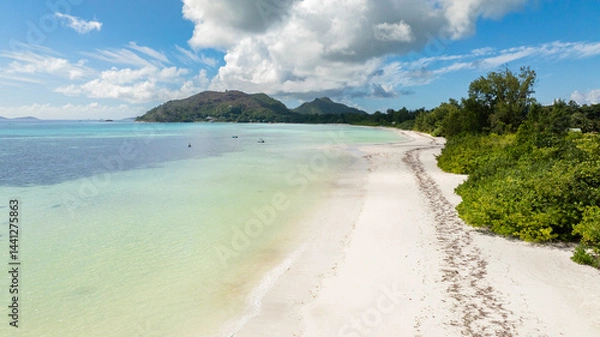 Fototapeta A pristine beach with soft white sand, lush palm trees, and boats floating in turquoise waters near a small island. Praslin, Seychelles.