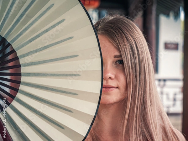 Fototapeta Young woman holds a decorative fan near her face in outdoor area with traditional architecture in the background.