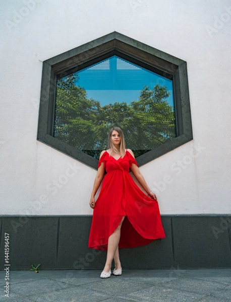 Fototapeta A woman stands elegantly in a flowing red dress, striking a pose in front of a unique hexagonal window.