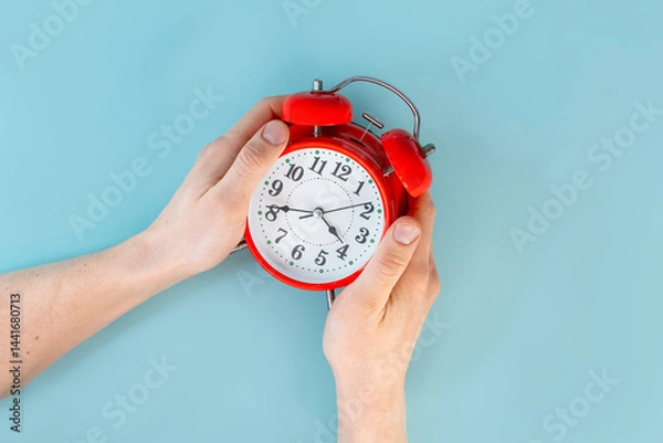 Obraz A man's hands are holding a red alarm clock on a blue table background. The concept of time, the place for the text. Close up, point of view shot.