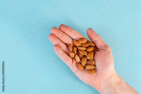 Obraz A man's hand holds a handful of almonds on a blue table background. The concept of healthy nutrition and vitamins. Close up, point of view shot.