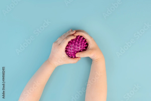 Obraz A little child's hands hold an orthopedic massage ball with spikes on a blue background. Close up, point of view shot. View from above.