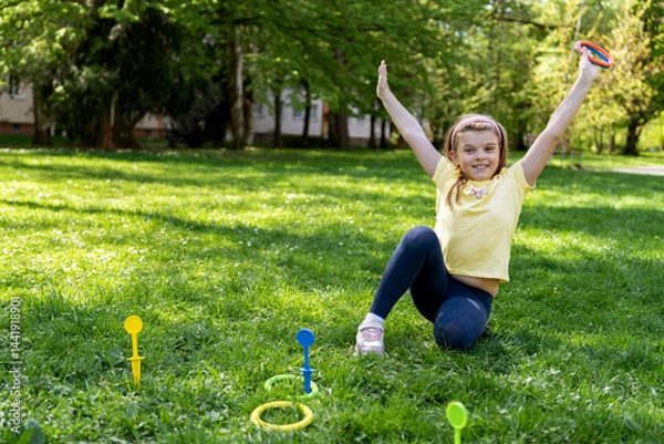 Fototapeta Joyful child sitting on the grass with raised arms, enjoying a summer day, playing ring toss in the park.