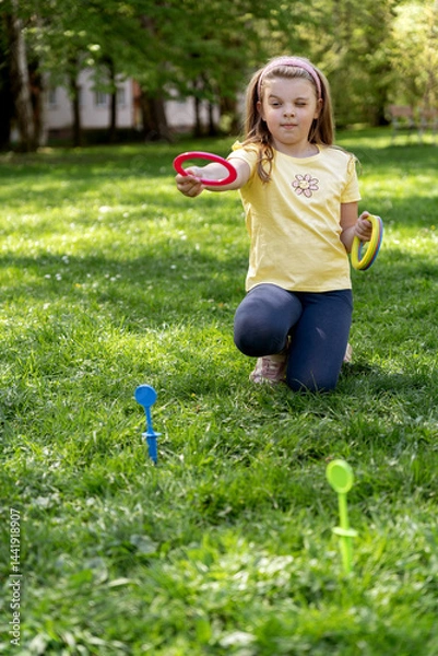 Fototapeta Focused little girl takes careful aim while playing colorful ring toss game outdoors on a bright sunny day. Fun children's games in the park.
