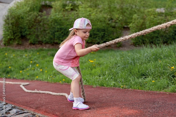 Fototapeta Preschool girl climbs a steep slope, holding on to a rope, during a physical education lesson at school or kindergarten. Side view. Sports games on the playground.