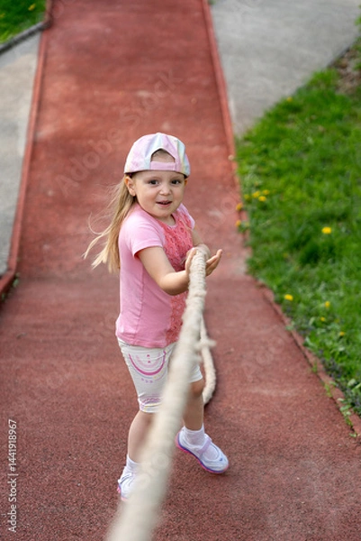 Fototapeta Little girl holds on to a rope to climb up a ledge. Physical education lesson at school or kindergarten.