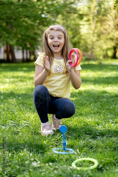 Fototapeta Happy girl celebrating a win in an outdoor ring toss game, smiling brightly on green grass in a sunny park.
