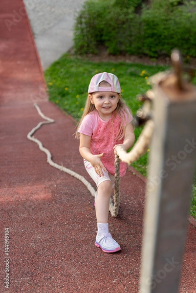 Fototapeta Little girl holds on to a rope to climb up a ledge. Physical education lesson at school or kindergarten. Games on a playground.