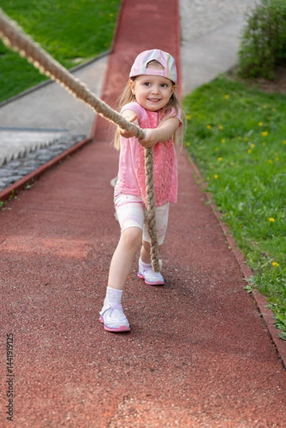 Fototapeta Preschool girl climbing up a steep slope holding onto a rope during a physical education lesson at school or kindergarten. Playing on a playground .