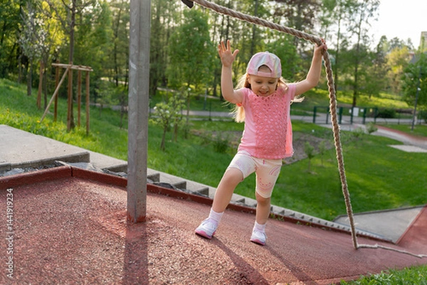 Fototapeta Happy little girl in pink clothes climbed a steep slope holding on to a rope. Physical education at school or kindergarten. Fun sports game on the playground.