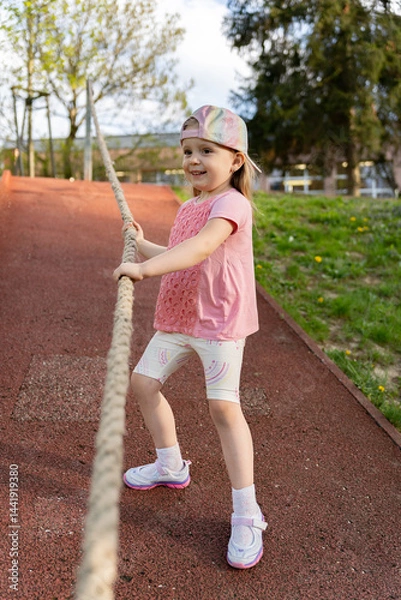 Fototapeta Little girl holds on to a rope to climb up a ledge.Fun sports game on the playground.
