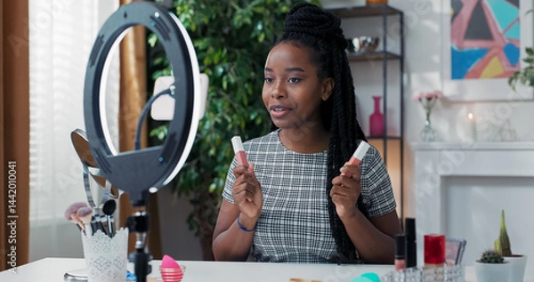 Fototapeta A female beauty blogger records a daily makeup tutorial using a ring light and a phone on a tripod, live streaming and comparing two shades of matte lipsticks.