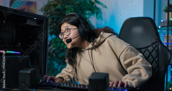 Fototapeta Focused female gamer wearing a microphone headset communicates with online rivals during a match, playing in a dark LED-lit room.