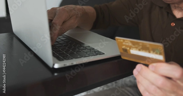 Fototapeta Hand of senior male buying online with credit card. Man using laptop and doing online transactions at home. Concept online shopping, technology and lifestyle, buying cryptocurrency