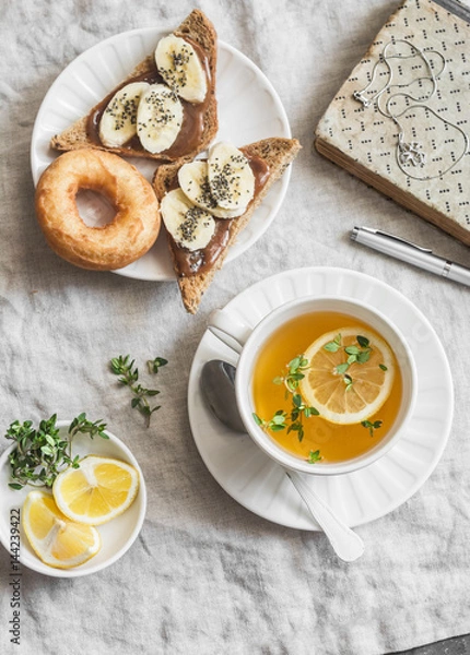 Fototapeta Green tea with lemon and thyme, banana sandwich with chia seeds, donuts on a light background, top view. Afternoon tea. Vintage style still life