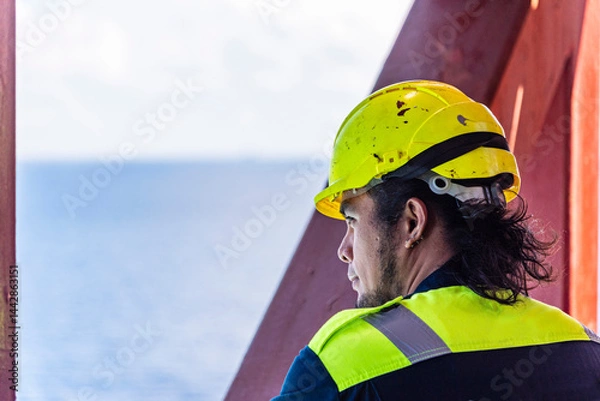 Fototapeta Portrait of a young Filipino officer in protective clothing and a yellow helmet, standing aboard a container ship - his eyes focused ahead, reflecting both duty and quiet determination.