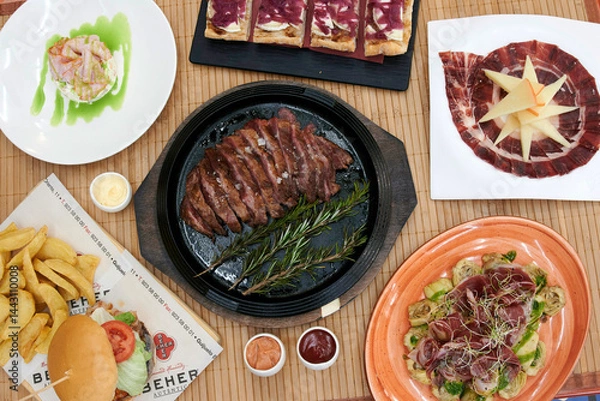 Obraz A table with a variety of food including a burger, a sandwich, and a salad. There are also plates of meat and vegetables